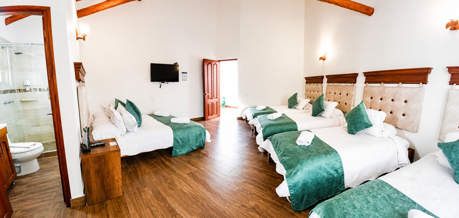 A hotel room with a wooden floor, a TV mounted on the wall, and a bed with green bedding.