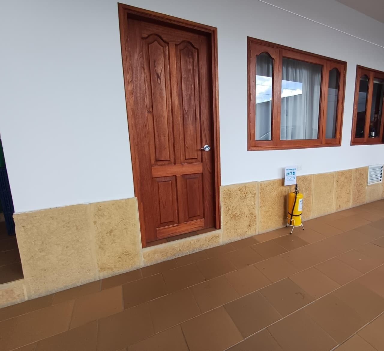 Un pasillo con una puerta de madera, ventanas con cortinas y un extintor.