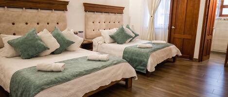 Habitación de hotel con dos camas, cabeceros de madera, una mesita de noche, una puerta de madera y una ventana con cortinas.