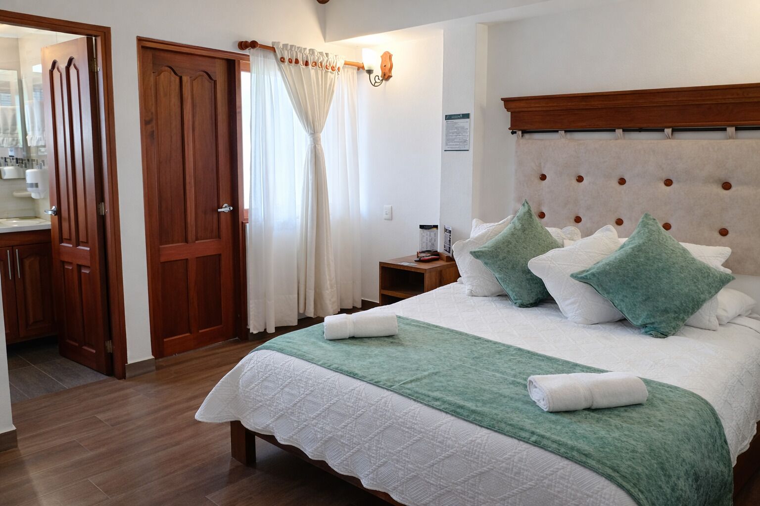 Una habitación de hotel con una cama, una puerta de madera, una mesita de noche y una ventana con cortinas.