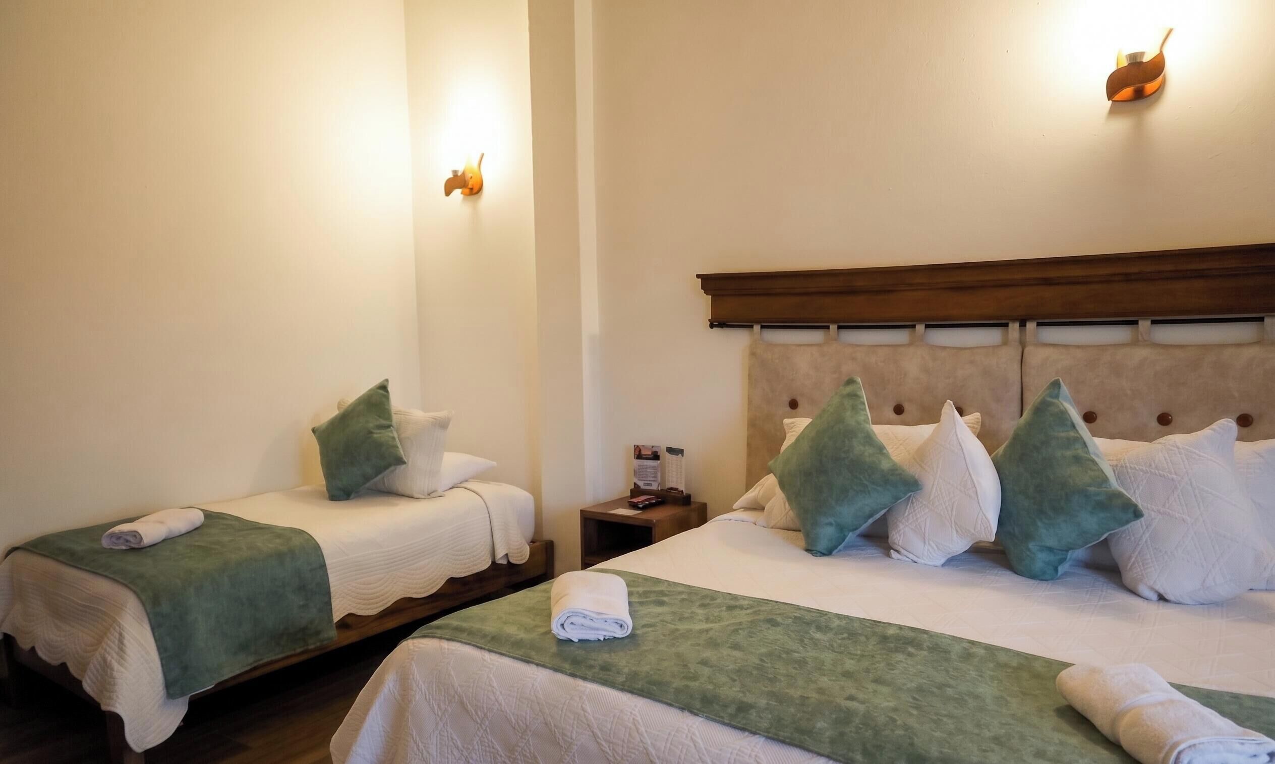 A hotel room with two beds, each with a green bedspread and white linens, a wooden headboard, and a nightstand with a lamp.