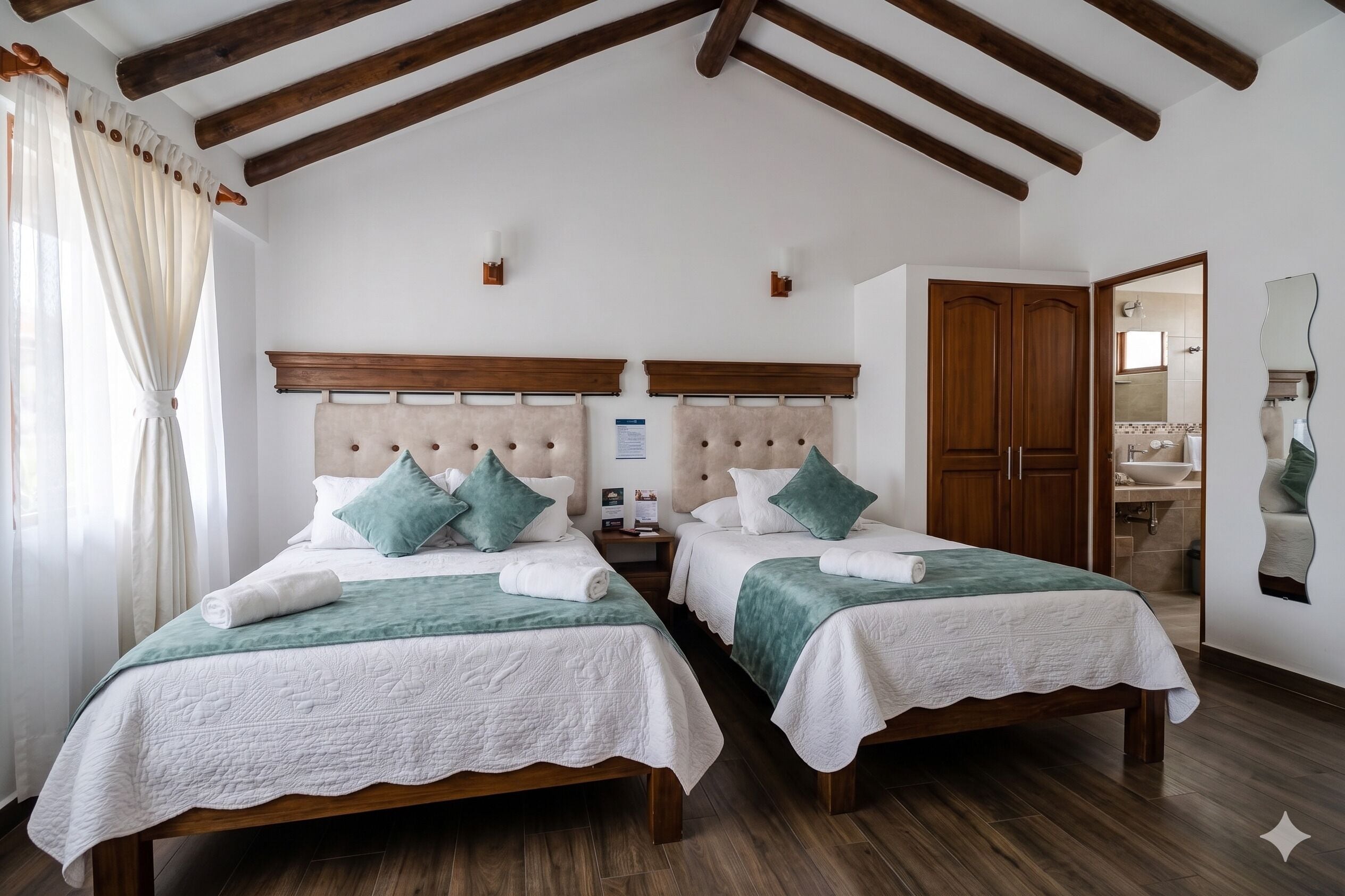 Habitación doble con dos camas, cabeceros de madera, techo de madera y un baño visible a través de una puerta abierta, además de un espejo en la pared.