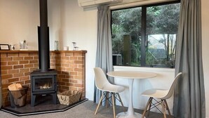 A room with a brick fireplace, a round table with two chairs, and a large window with curtains. - Bushside Cottage - Akaroa Holiday Home (Akaroa)