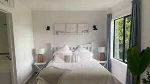 A bedroom with a bed, two bedside tables, a window with curtains, and wall art. - Bushside Cottage - Akaroa Holiday Home (Akaroa)