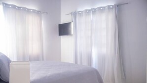 Une chambre à coucher avec un lit, des rideaux, un téléviseur à écran plat fixé au mur et un sol carrelé.