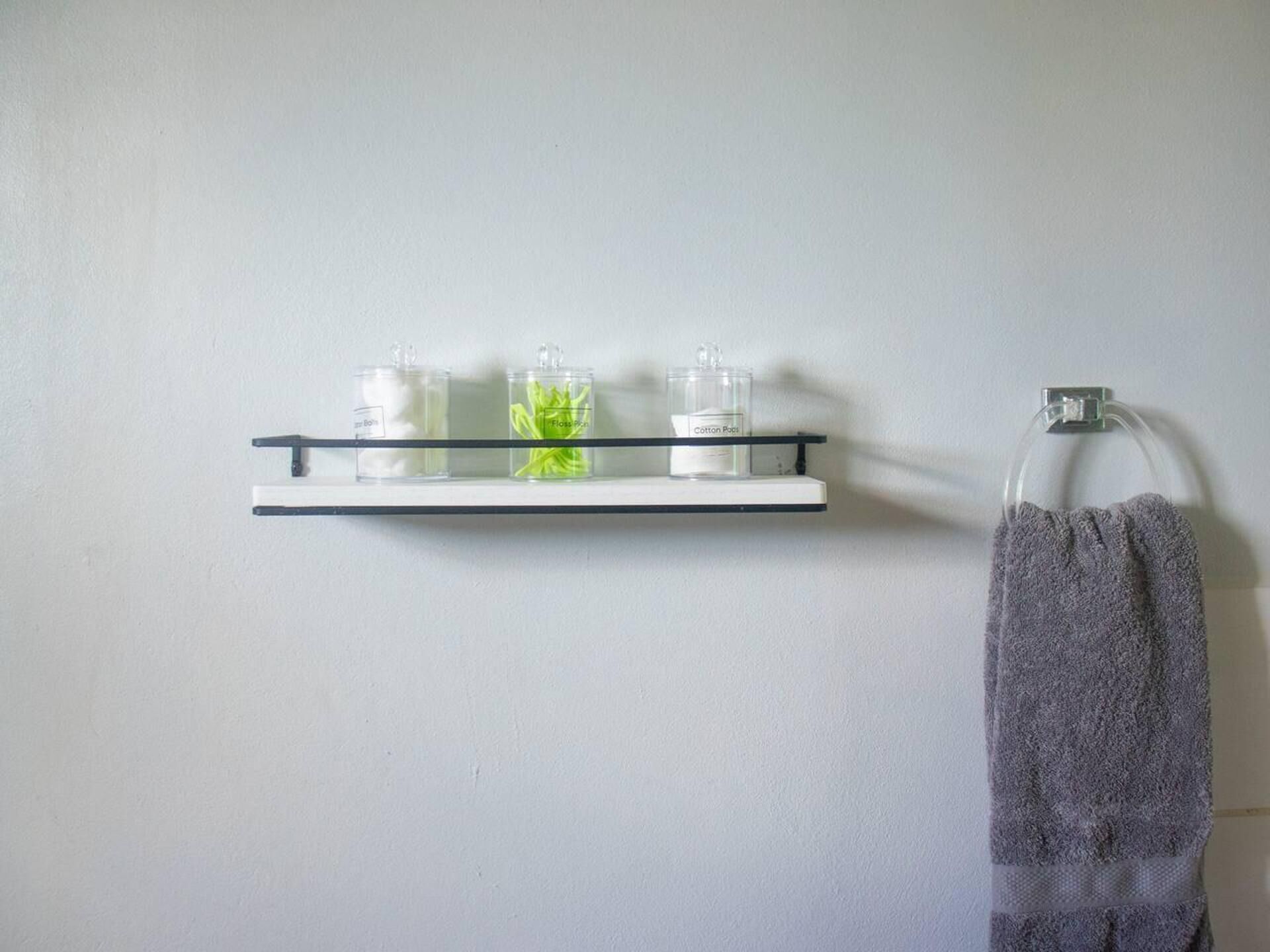 Un baño con pared blanca, un estante con tres recipientes y una toalla colgada en un gancho.