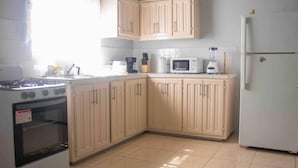 Une cuisine avec des meubles de rangement en bois clair, une cuisinière à gaz, un four à micro-ondes, un mixeur et un réfrigérateur.