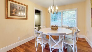 A dining area with a round table, white chairs, a chandelier, and a window with blinds.