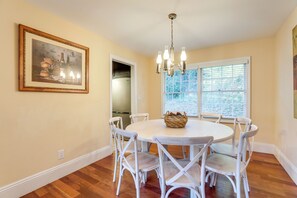 A dining area with a round table, white chairs, a chandelier, and a window with blinds.