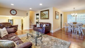 A spacious living room with leather sofas, a glass coffee table, and a dining area with white chairs and a chandelier.