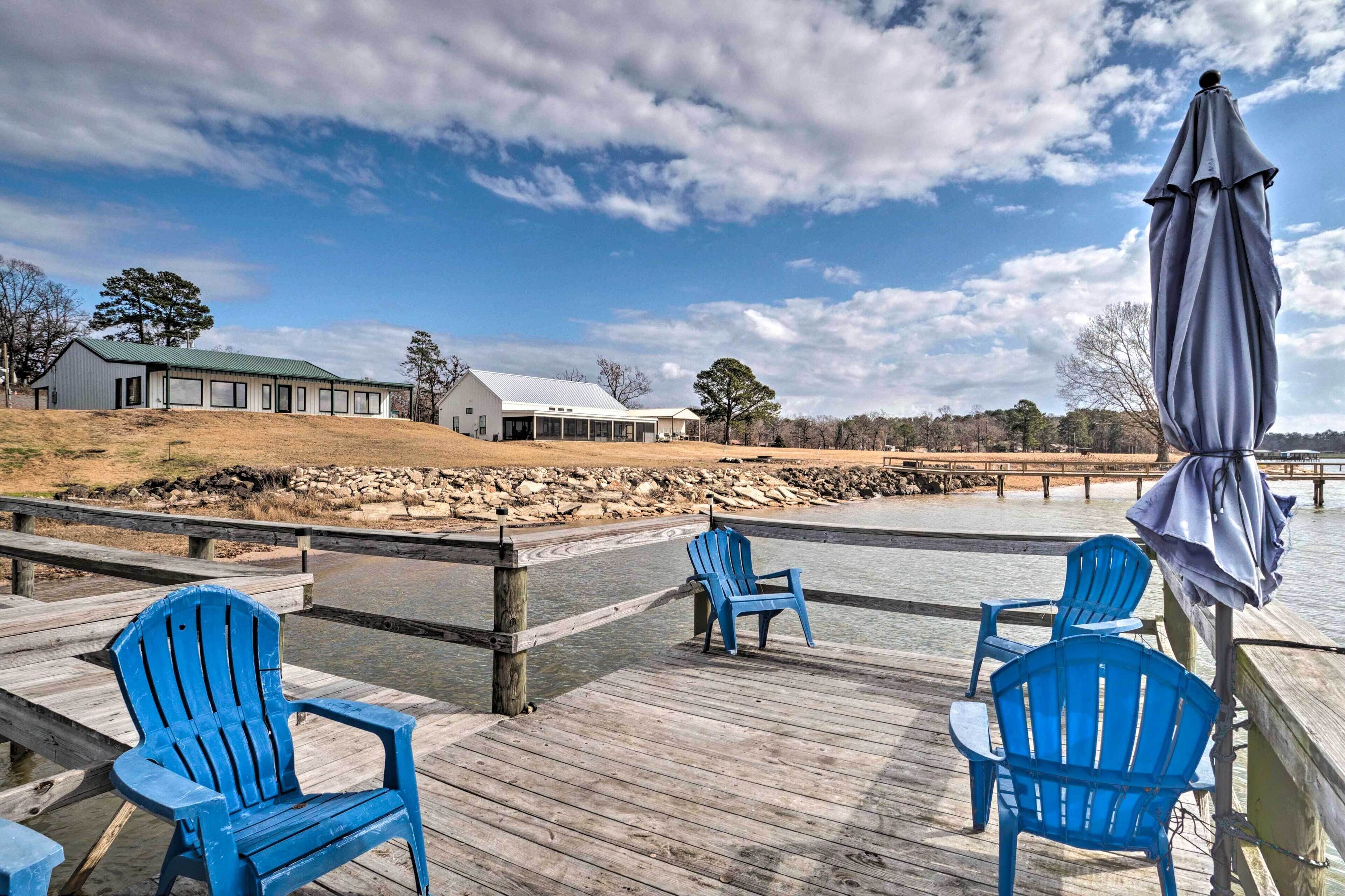 A wooden deck with blue chairs and a covered umbrella, overlooking a calm body of water and a building with a green roof.