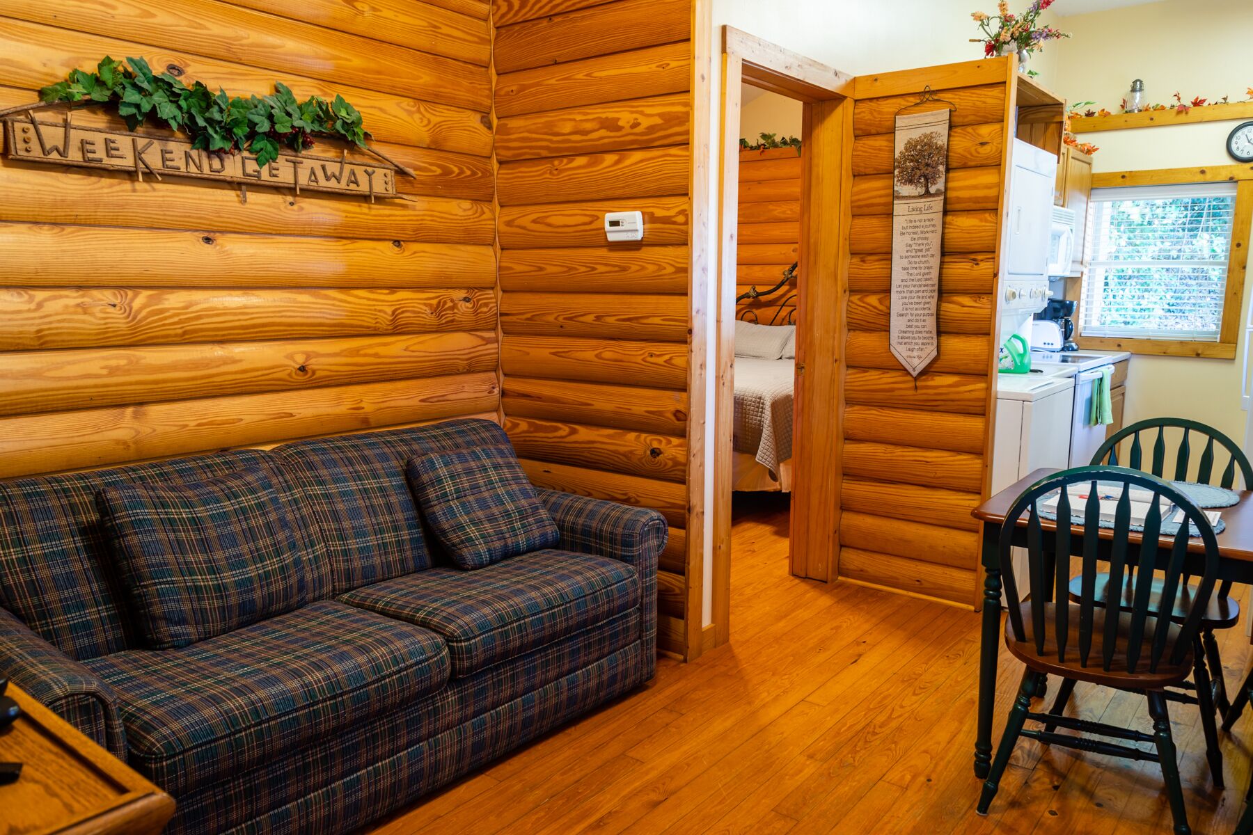 A cozy log cabin interior with a plaid sofa, a dining area with two chairs, a kitchen visible through a window, and a sign on the wall reading "Weekend Getaway".