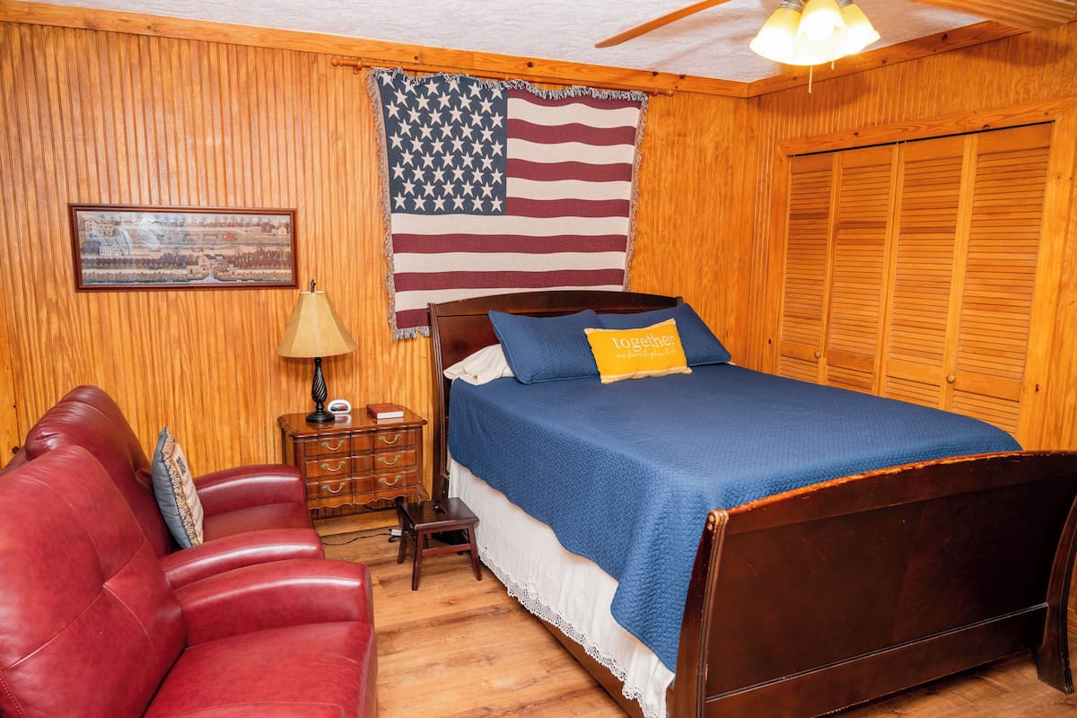 A bedroom with a wooden floor, a bed with a blue bedspread and a yellow pillow, a red leather couch, a wooden nightstand with a lamp, and an American flag on the wall.