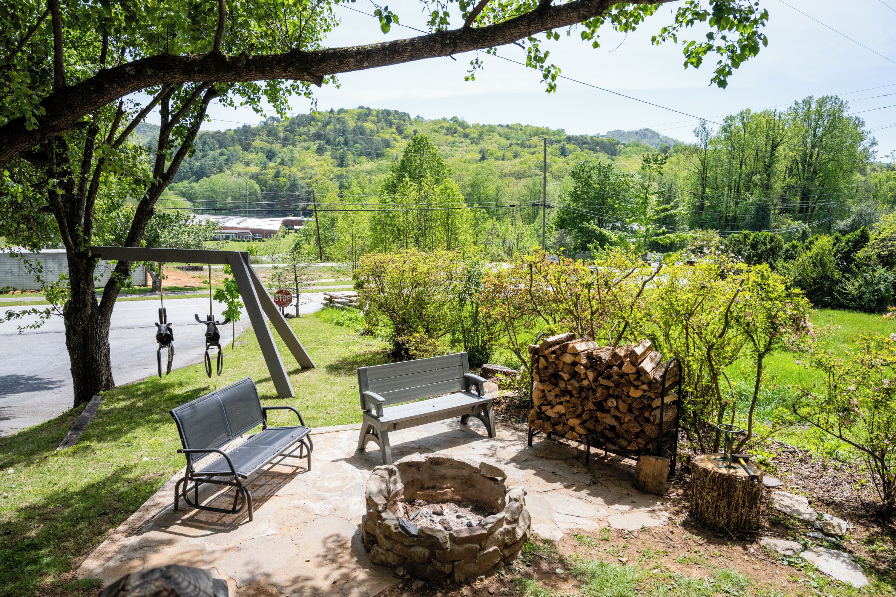 Un espace extérieur aménagé avec des bancs et un foyer, entouré d’arbres et offrant une vue sur un paysage vallonné.