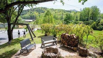 Un espace extérieur aménagé avec des bancs et un foyer, entouré d’arbres et offrant une vue sur un paysage vallonné.