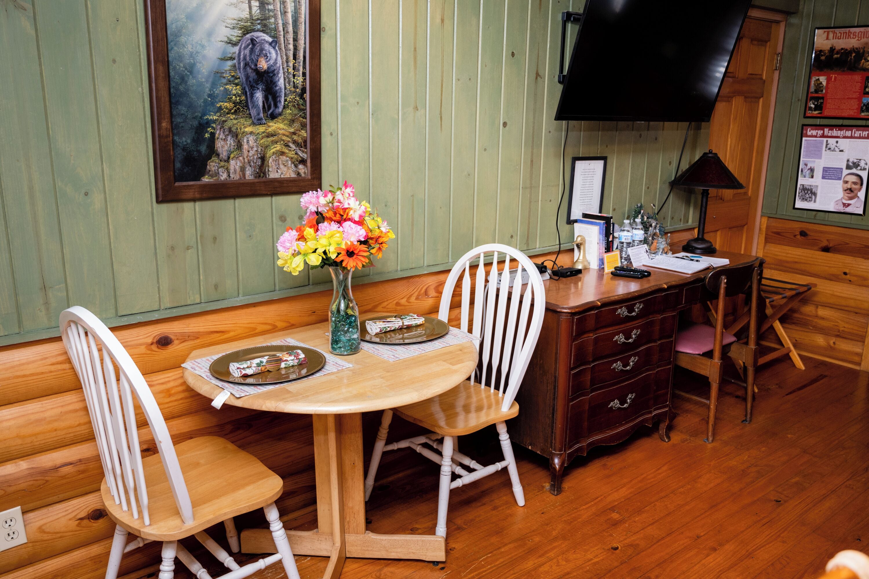 A wooden dining table with white chairs, a vase of flowers, and a painting of a bear on the wall.