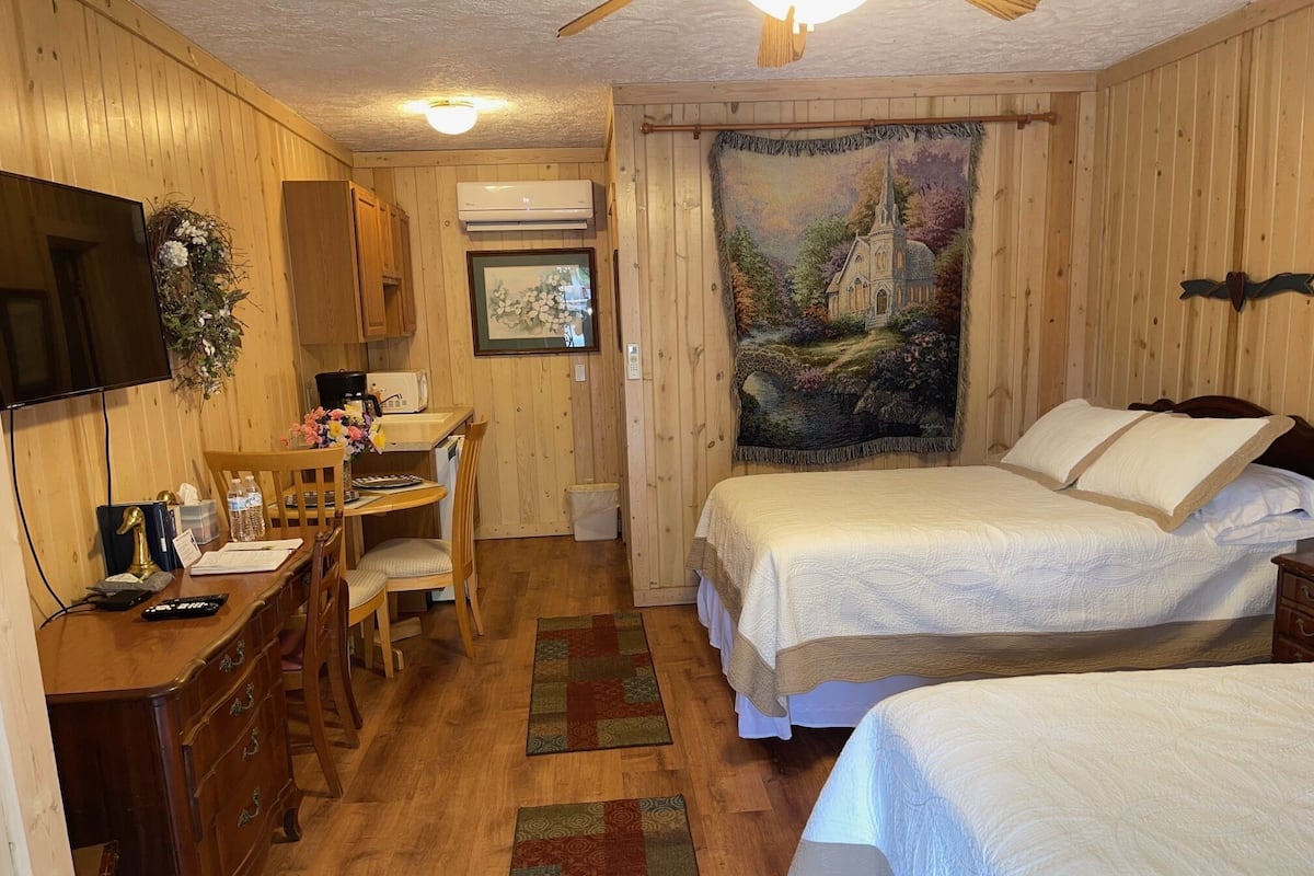 A cozy room with wooden walls, a bed with white bedding, a desk with a chair, a television, and a painting on the wall.