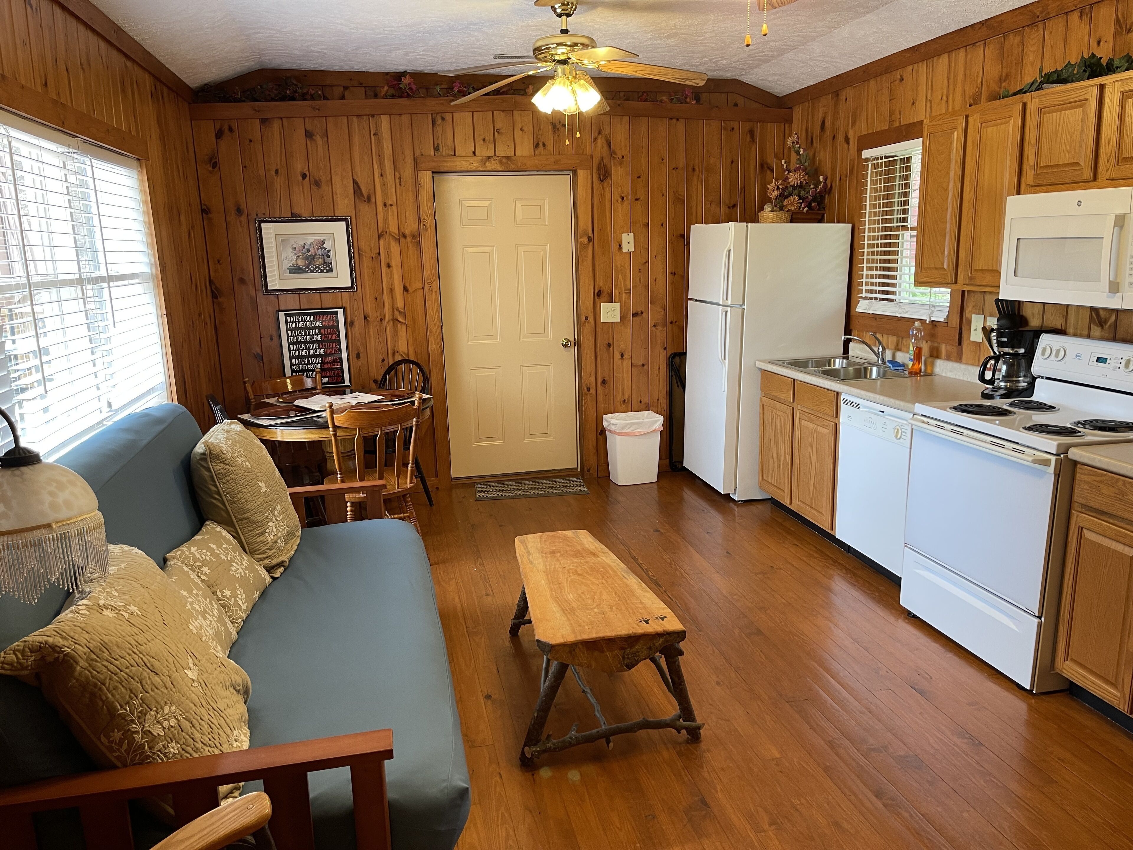 A cozy room with a wooden floor, a blue sofa, a wooden bench, a dining table with chairs, a white refrigerator, a microwave, a stove, and a ceiling fan.