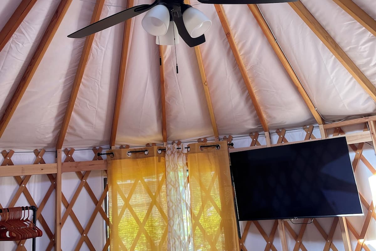 A room with a ceiling fan, a TV mounted on the wall, and a window with curtains.