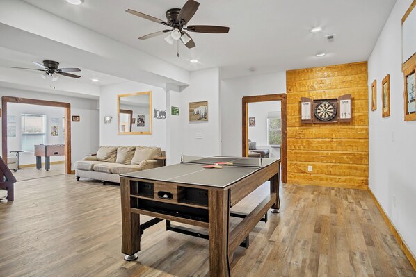 A spacious living room with a ping pong table, ceiling fans, and a dartboard on a wooden wall. - 3 King Bedrooms with Hot Tub Game Room Fire Pit Pool Table Putting Room (Townsend)