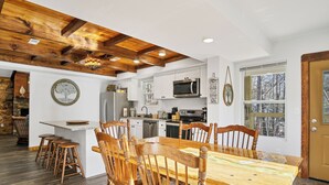 A spacious kitchen with wooden beams, a dining area with a large wooden table and chairs, and a view of trees through a window. - 3 King Bedrooms with Hot Tub Game Room Fire Pit Pool Table Putting Room (Townsend)