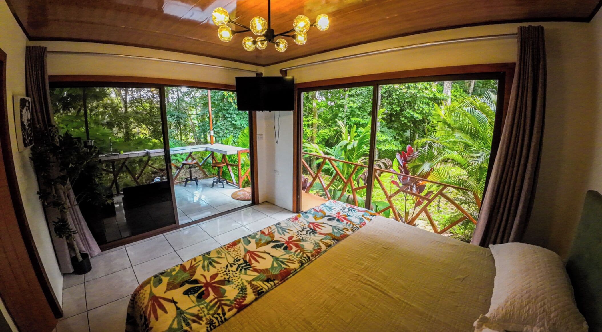 A bedroom with a large bed, a TV, and a balcony with a view of lush greenery.