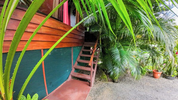 Un escalier en bois mène à un bâtiment à la façade verte et brune, entouré d’une végétation tropicale luxuriante.