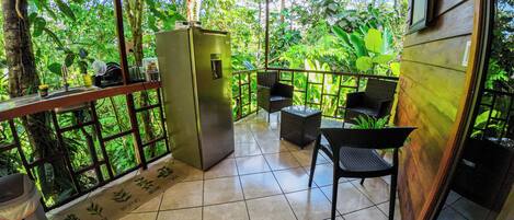 Un balcón con refrigerador, sillas y una mesa, rodeado de exuberante vegetación.