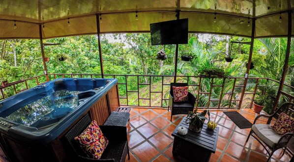 A covered outdoor area with a hot tub, seating, and potted plants.