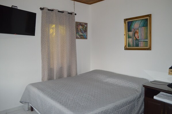 Un dormitorio con una cama, televisión montada en la pared, un cuadro y una cortina.