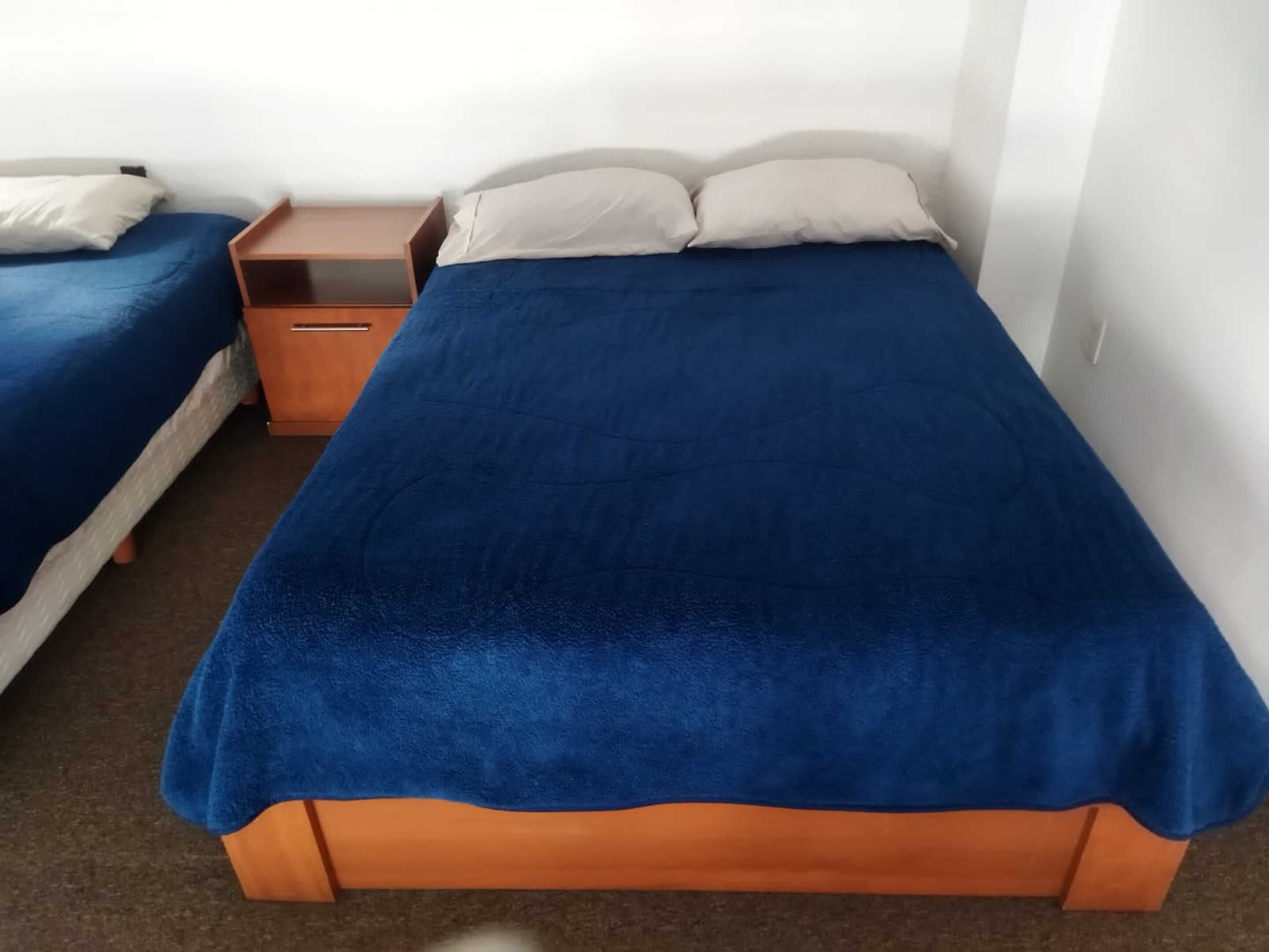 A single bed with a blue blanket and two pillows, a wooden nightstand with a drawer, and a white wall.