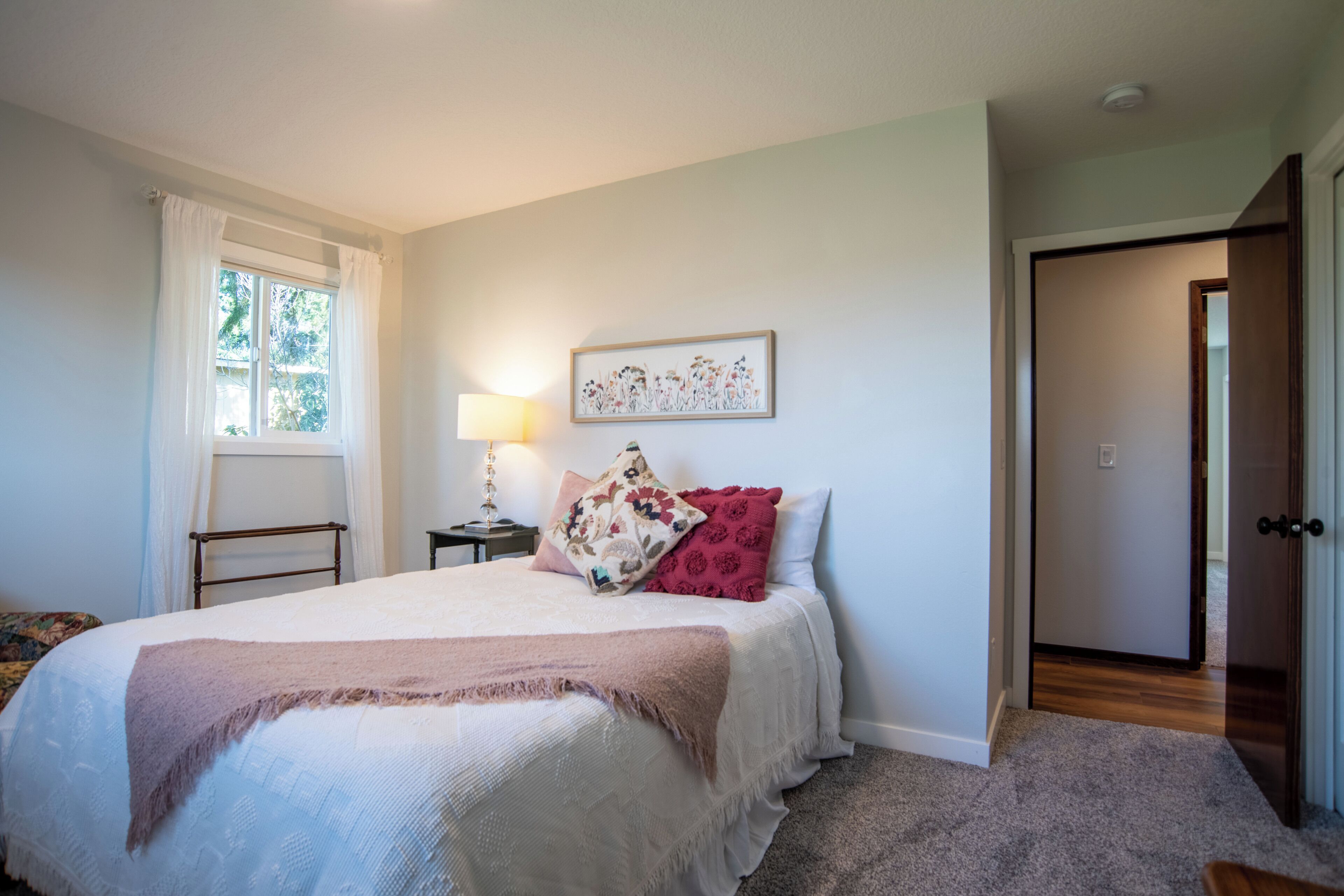 A bedroom with a bed, a bedside table with a lamp, a window with curtains, a door, and a wall with a framed picture.