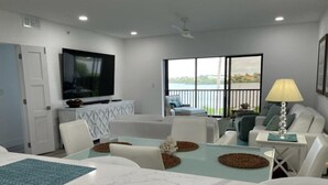 Een moderne woonkamer met een glazen eettafel, witte banken, een flatscreen-televisie en een balkon met uitzicht op het water.