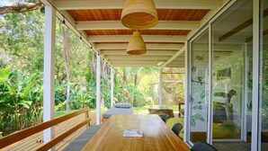 Mesa de madeira com um laptop, rodeada por cadeiras, num quarto com paredes de vidro e vista para o verde.