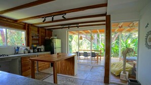 Cozinha com vigas de madeira, área de refeições com mesa e cadeiras, e vista para o verde lá fora.