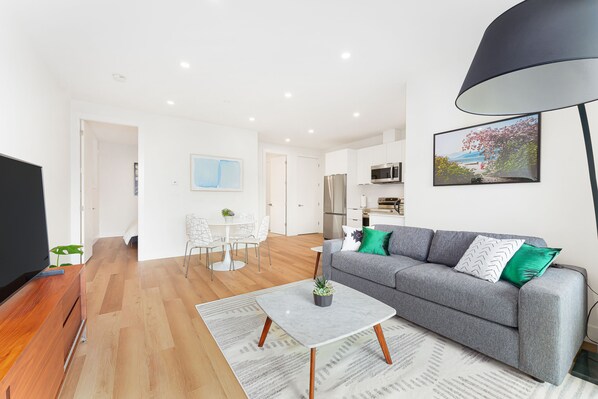 Ein modernes Wohnzimmer mit grauem Sofa, einem Couchtisch und einem Fernseher. Es gibt eine Essecke mit Tisch und Stühlen sowie eine Küche im Hintergrund.