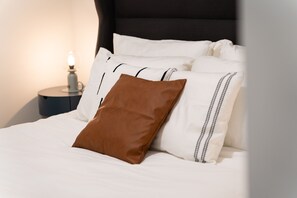 Ein ordentlich bezogenes Bett mit weißen und braunen Kissen, einem dunklen Kopfteil und einem Nachttisch mit einer Lampe.