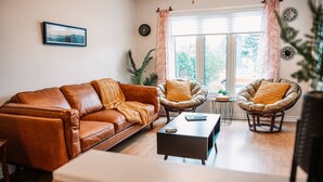 Een woonkamer met een bruine leren bank, twee rieten fauteuils, een salontafel en een raam met gordijnen.