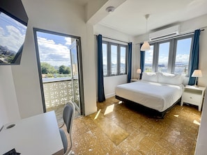 A hotel room with a bed, a desk, and a chair. There are windows with curtains and an air conditioning unit. - Bahia Urbana Hotel (San Juan)
