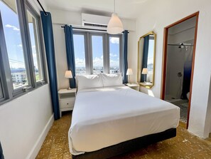 Een hotelkamer met een groot bed, twee nachtkastjes met lampen, een spiegel en uitzicht op de stad door grote ramen.