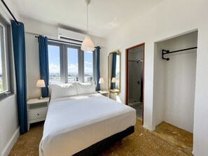 Een hotelkamer met een bed, nachtkastje, lamp, grote spiegel, raam met gordijnen en een douchehoek.