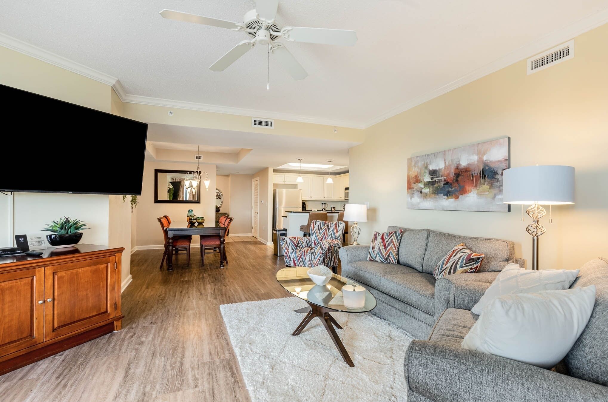 A living room with a sofa, coffee table, and TV. There is a dining area with chairs and a table. The room has wooden flooring and a ceiling fan.