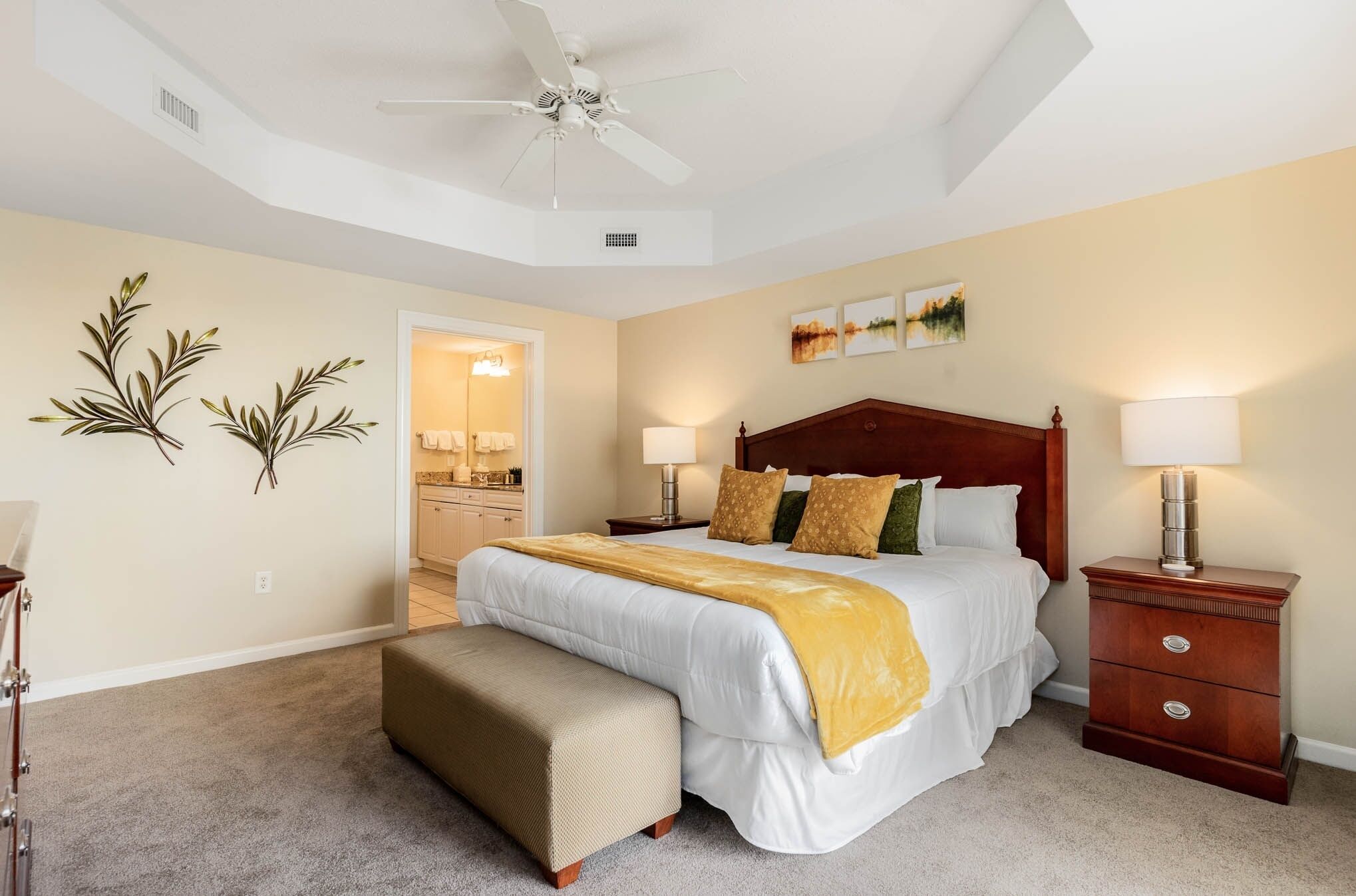 A bedroom with a bed, bedside tables, a bench, a ceiling fan, and artwork on the wall.