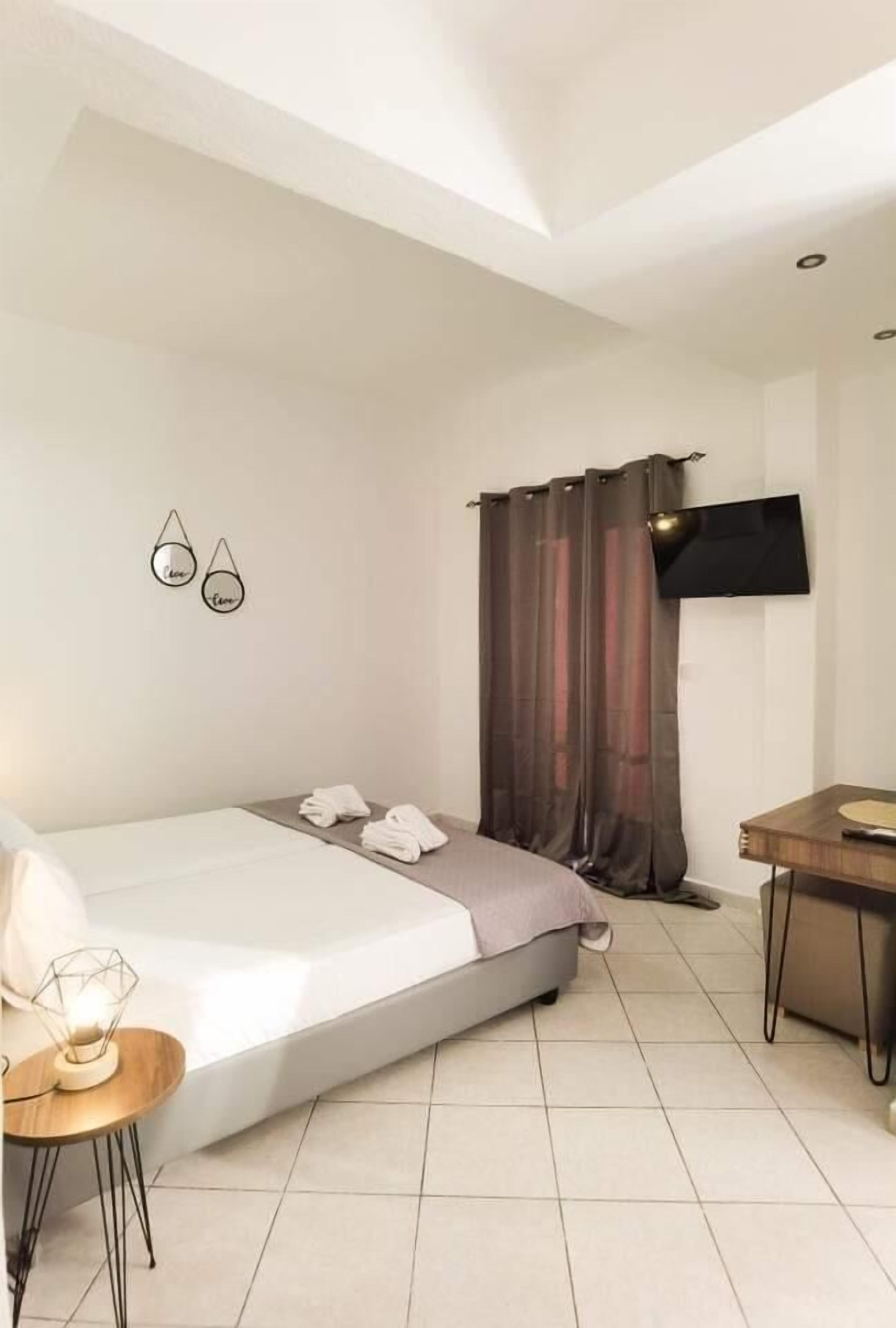 A modern hotel room with a bed, a small table with a lamp, a TV mounted on the wall, and a window with curtains.