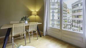 Un coin repas avec une table en bois et des chaises en osier, une plante en pot, et une vue sur des bâtiments historiques par une grande fenêtre.