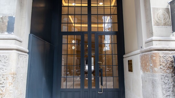 Une entrée à encadrement noir avec une porte en verre, un intérieur visible grâce à l’éclairage, et un mur en pierre orné d’une plaque.