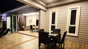 Un espace extérieur aménagé pour prendre les repas, avec une table et des chaises, une porte-fenêtre coulissante donnant sur une pièce et une fenêtre avec des rideaux.