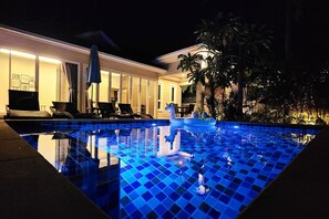 A nighttime view of a swimming pool with blue tiles, surrounded by a well-lit patio area with lounge chairs and umbrellas. - Ao Nang private pool villa near beach! (Ao Nang)