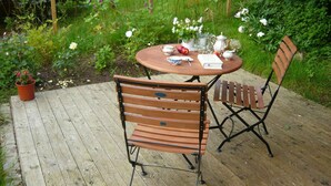 A wooden deck with a round table and two chairs, surrounded by lush greenery and flowering plants. - 3-room vacation apartment in the Allgäu with garden and wood-burning stove (Leutkirch im Allgäu)