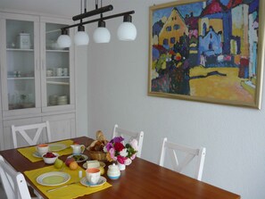 Ein Esstisch ist gedeckt mit Tellern, Schüsseln, einer Kanne und einem Brotkorb. Darüber an der Wand hängt ein Gemälde.
